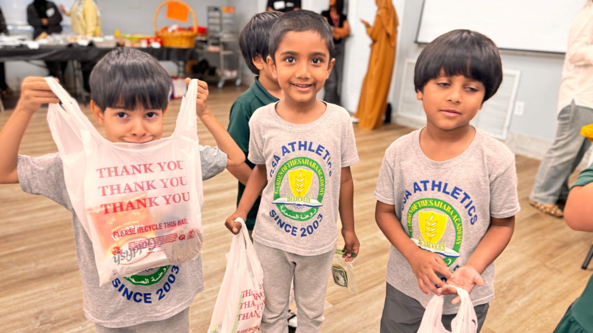 Three young students hold up their bags of goodies, one peeking over a Thank You bag