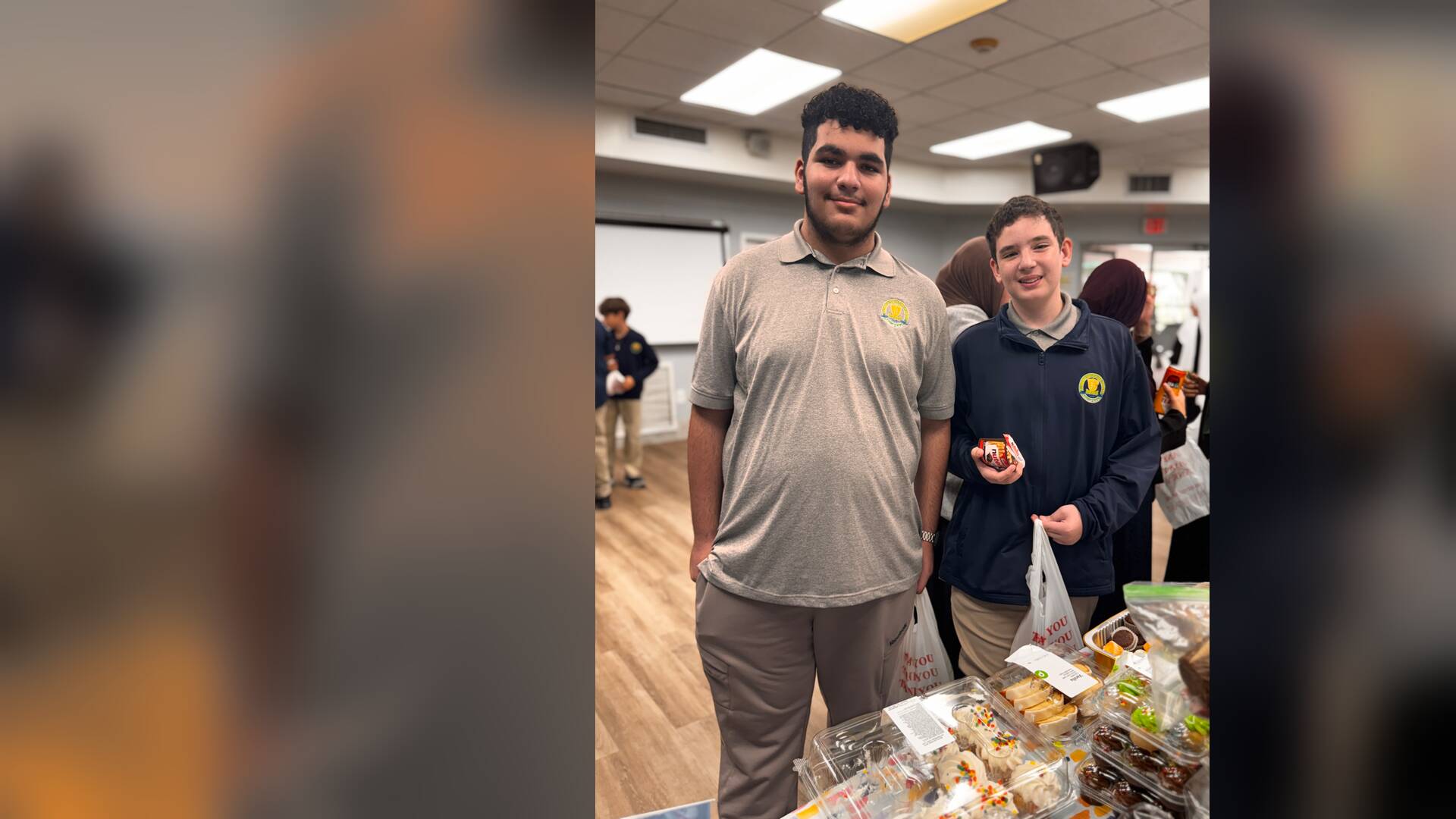 Two older students smile beside a table of frosted cupcakes, one holding a treat