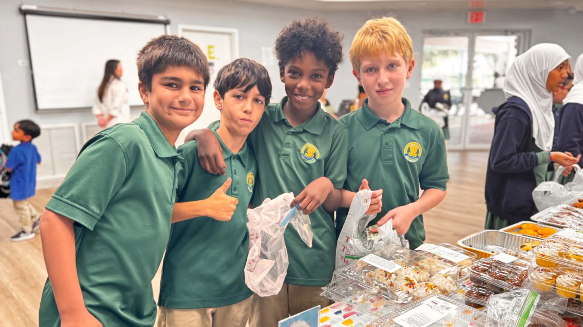 Four students in green polos give a thumbs-up at the dessert table