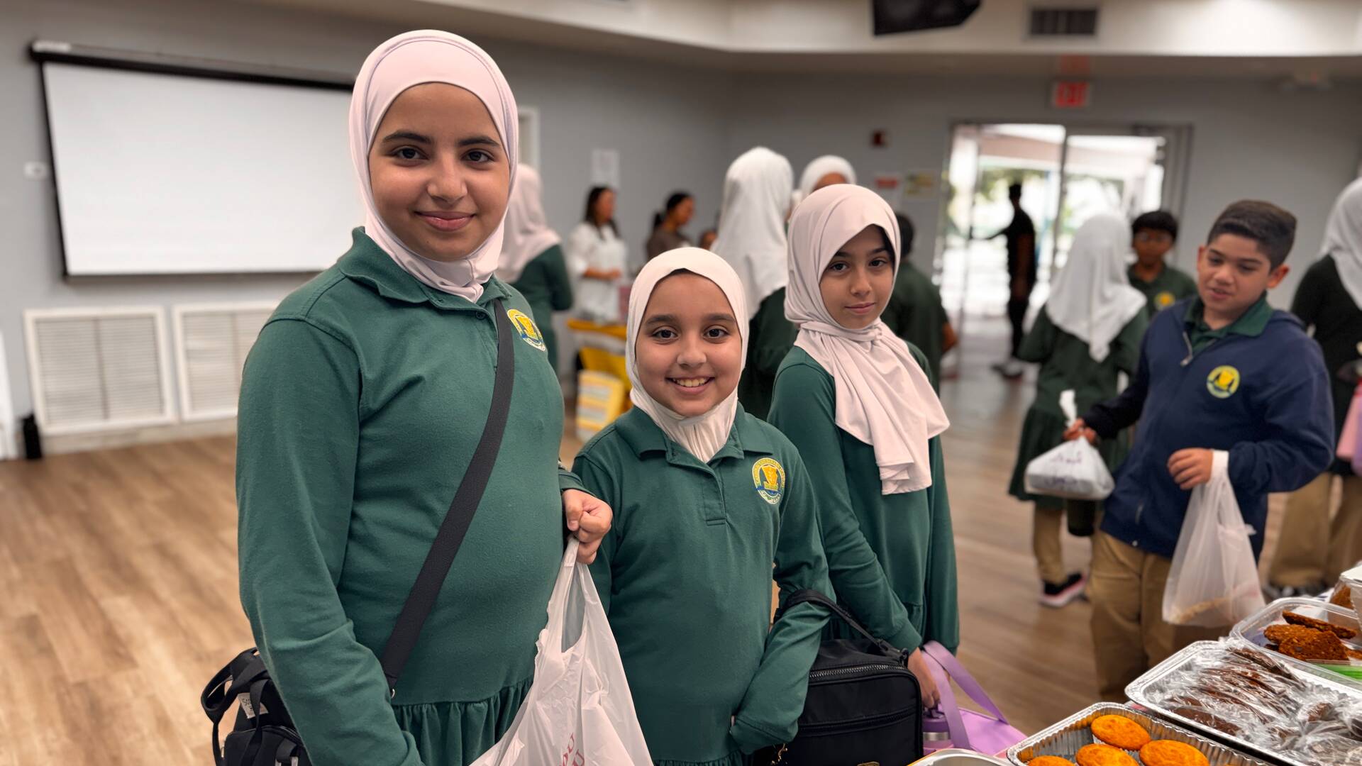 Three smiling students in green uniforms stop by the dessert table
