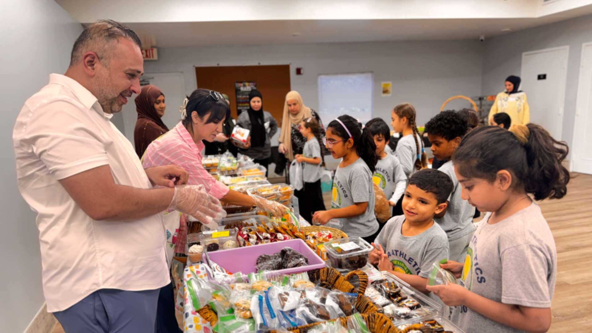 A gloved volunteer helps young students at a long table of sweets