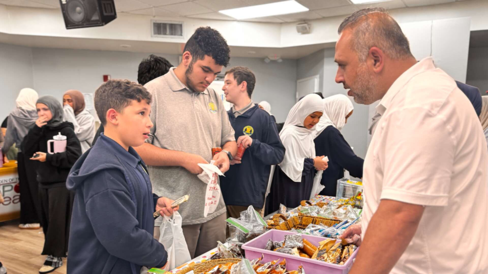 A volunteer helps an older student at a table of snacks and baked goods