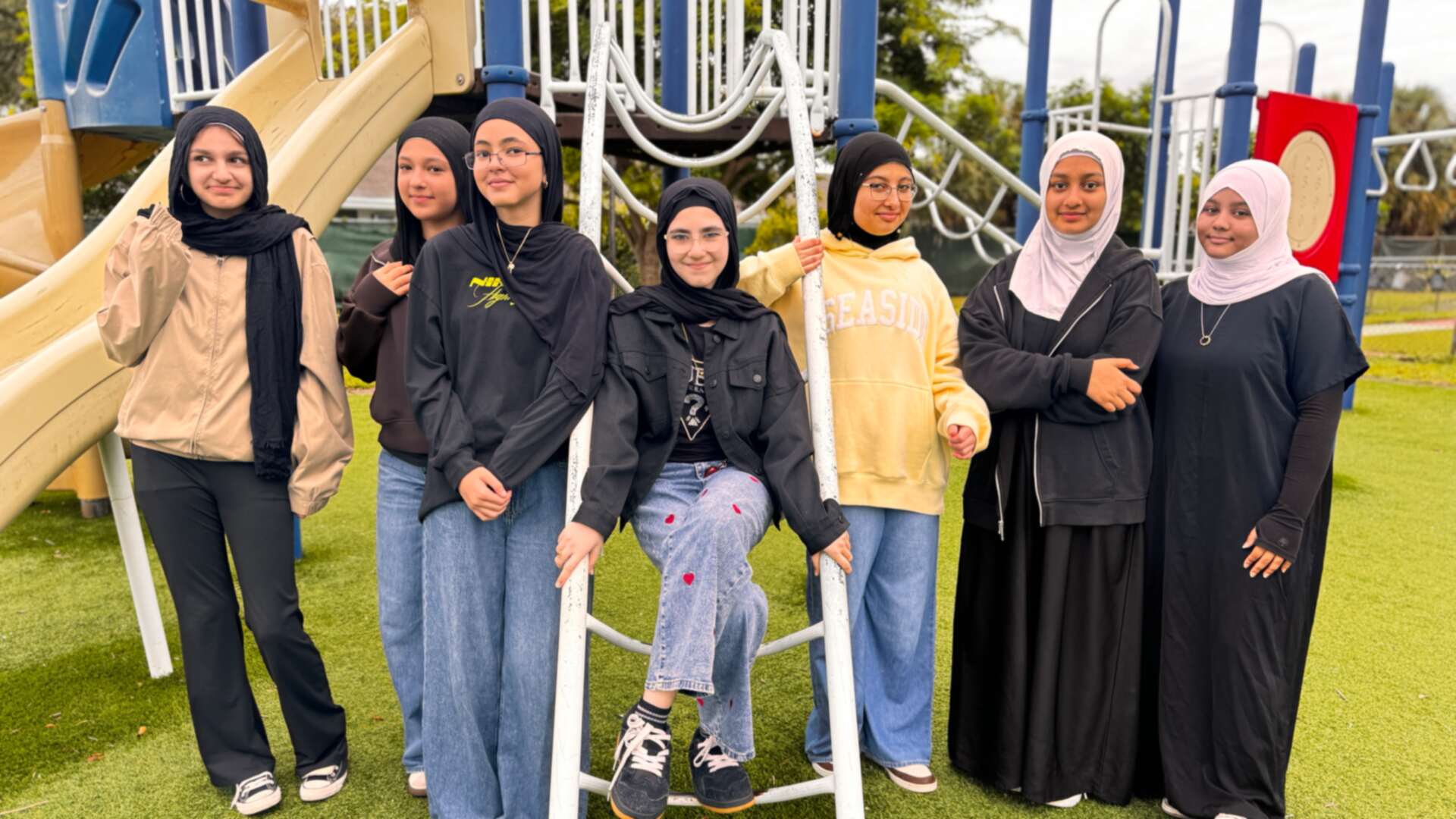 Friends in cozy hoodies and long dark dresses line up by the playground ladder on Dress Down Day