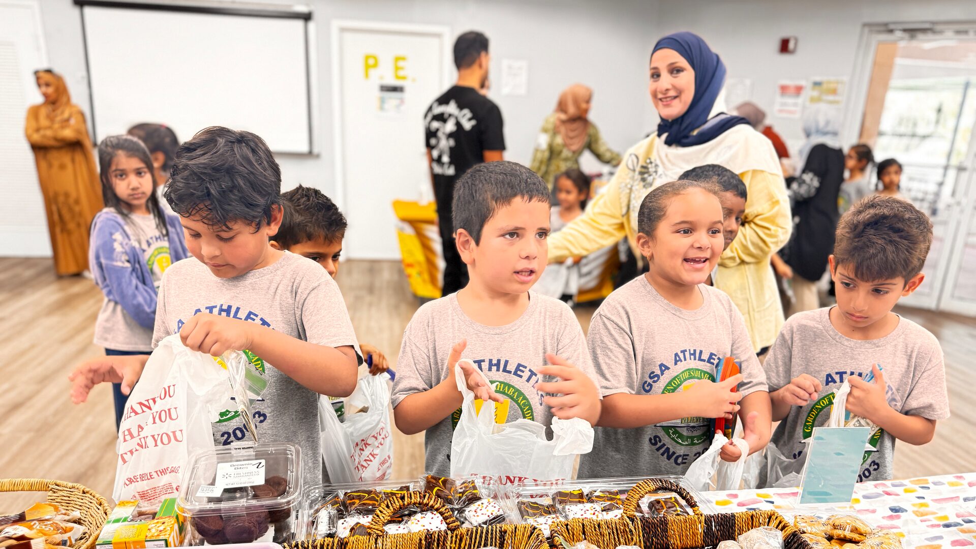 Young students hold bags at a table of wrapped treats while a parent volunteer smiles behind them.