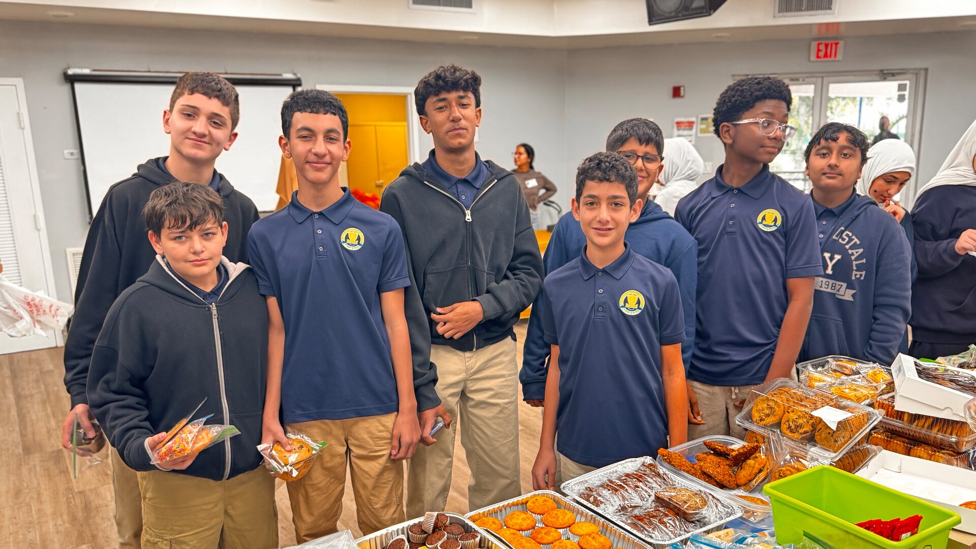 Students in navy school polos stand behind trays of muffins and cookies at the bake sale.