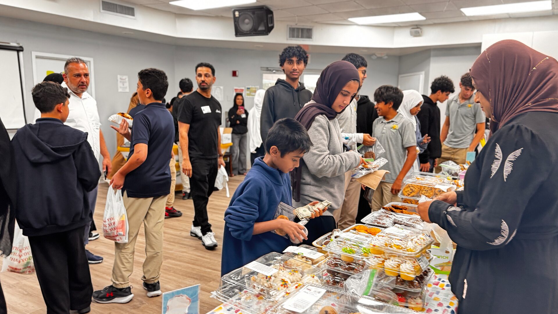 Students crowd around tables of baked goods at the bake sale while a volunteer serves them.