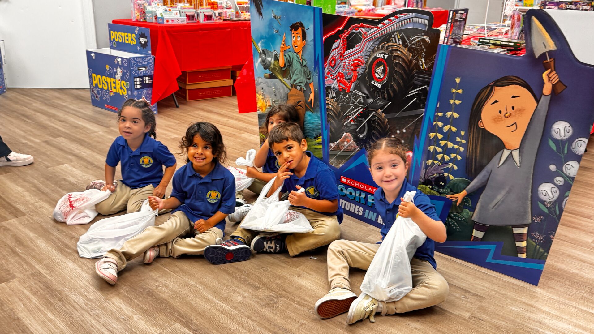 Young students in navy uniforms sit on the floor with their bags in front of a book fair display.