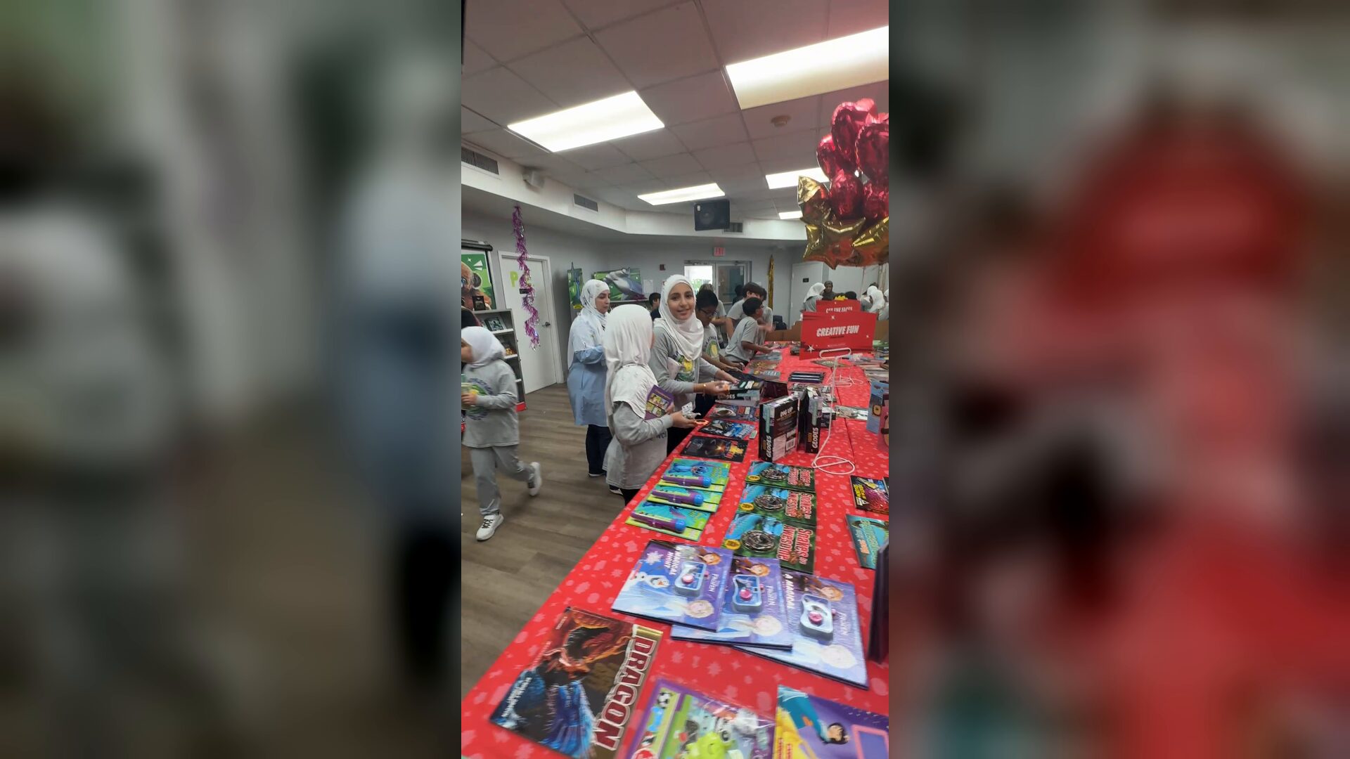 The camera moves along a red book fair table covered with books toward students browsing and smiling at the far end.