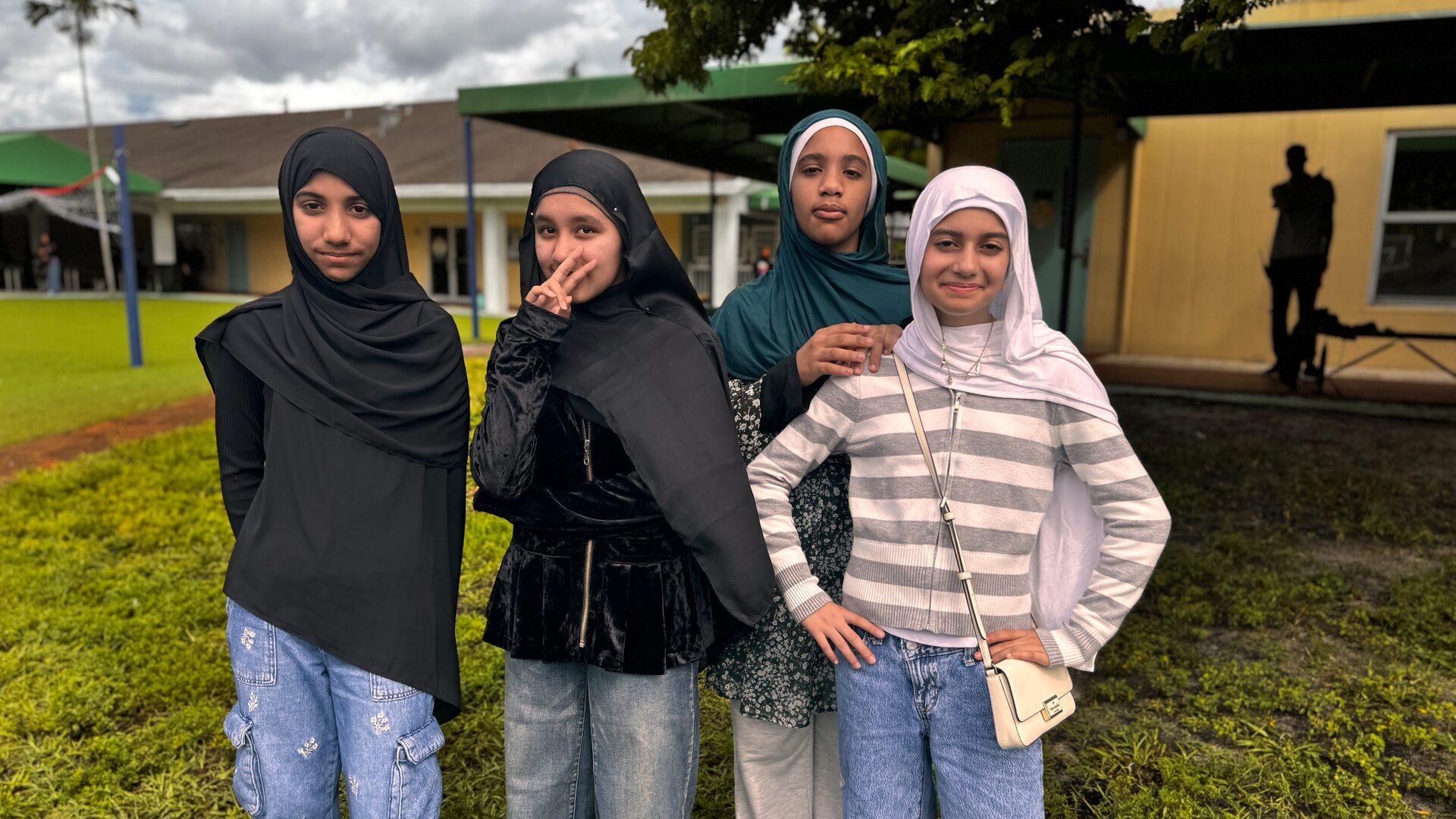 Four students in everyday clothes and hijabs pose together on the grass outside the school.
