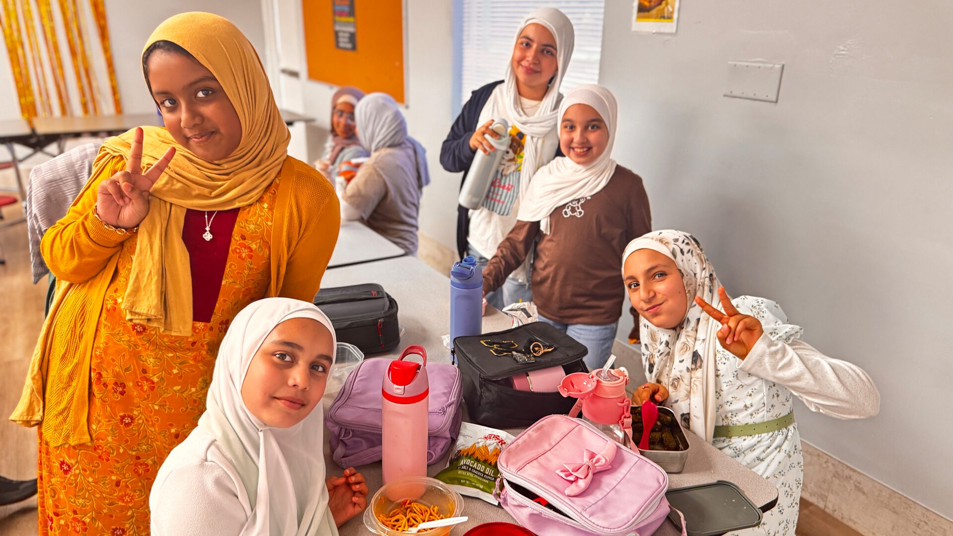Students in everyday clothes smile and make peace signs around a lunch table.