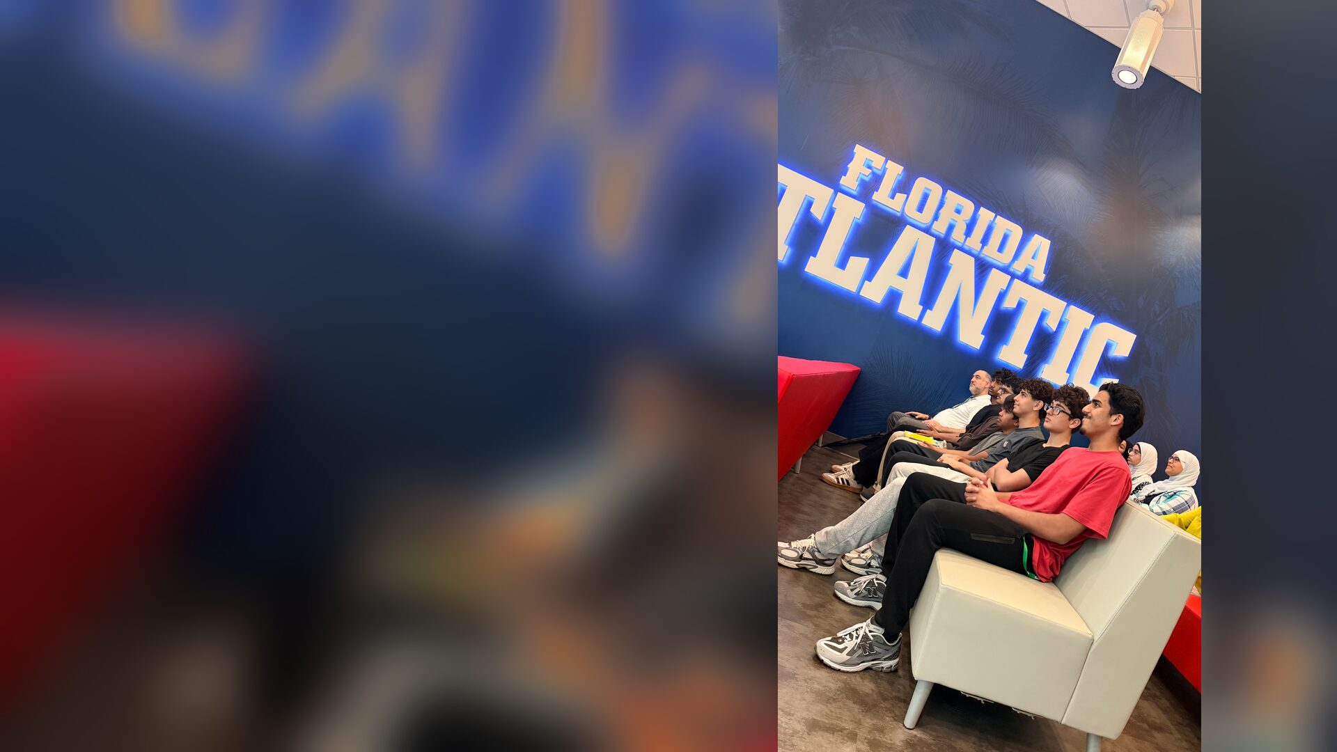 Students sit on a row of couches beneath the glowing FLORIDA ATLANTIC wall letters, listening to a presentation.