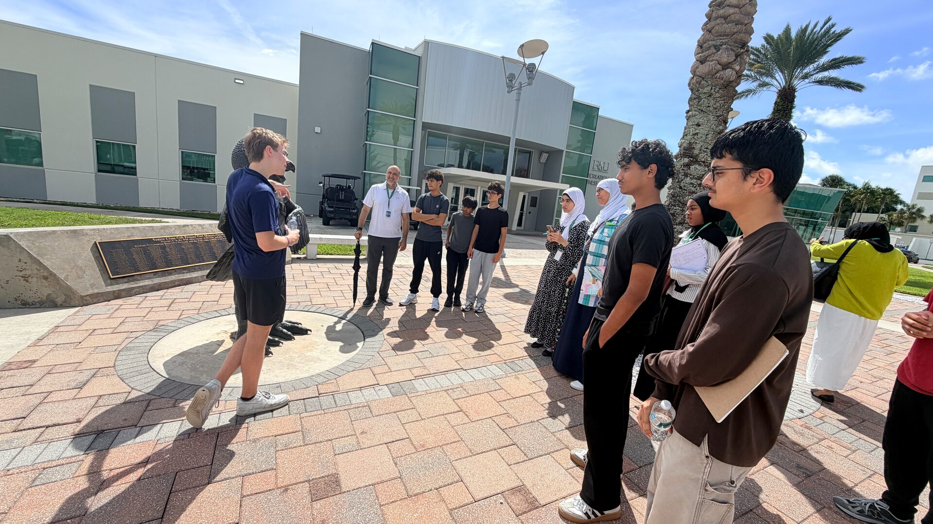 A campus tour guide speaks with a group of students in a sunny brick plaza.