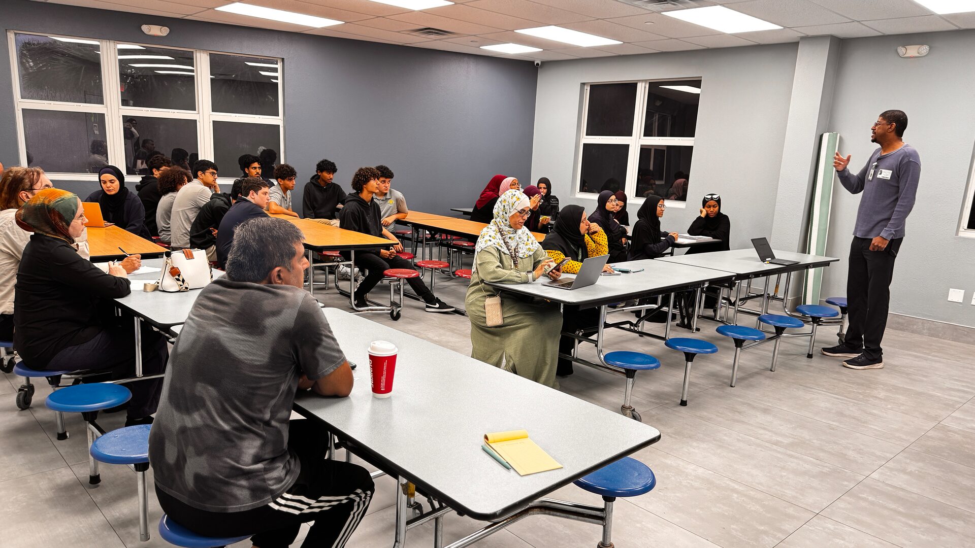 A presenter speaks to students and parents seated at long tables in the school cafeteria.