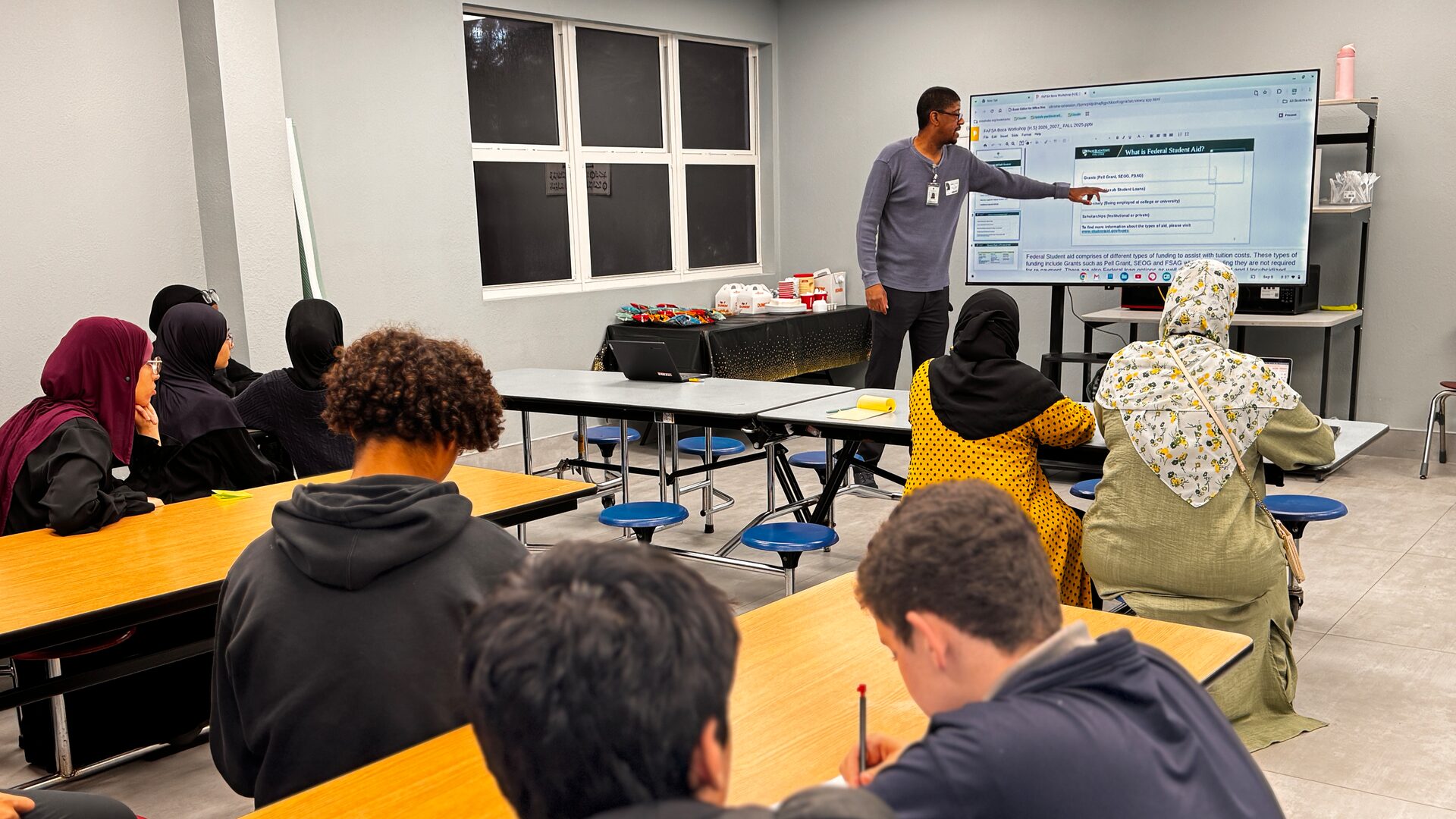 Students and parents sit at cafeteria tables facing a presenter beside a screen showing a federal student aid slide.