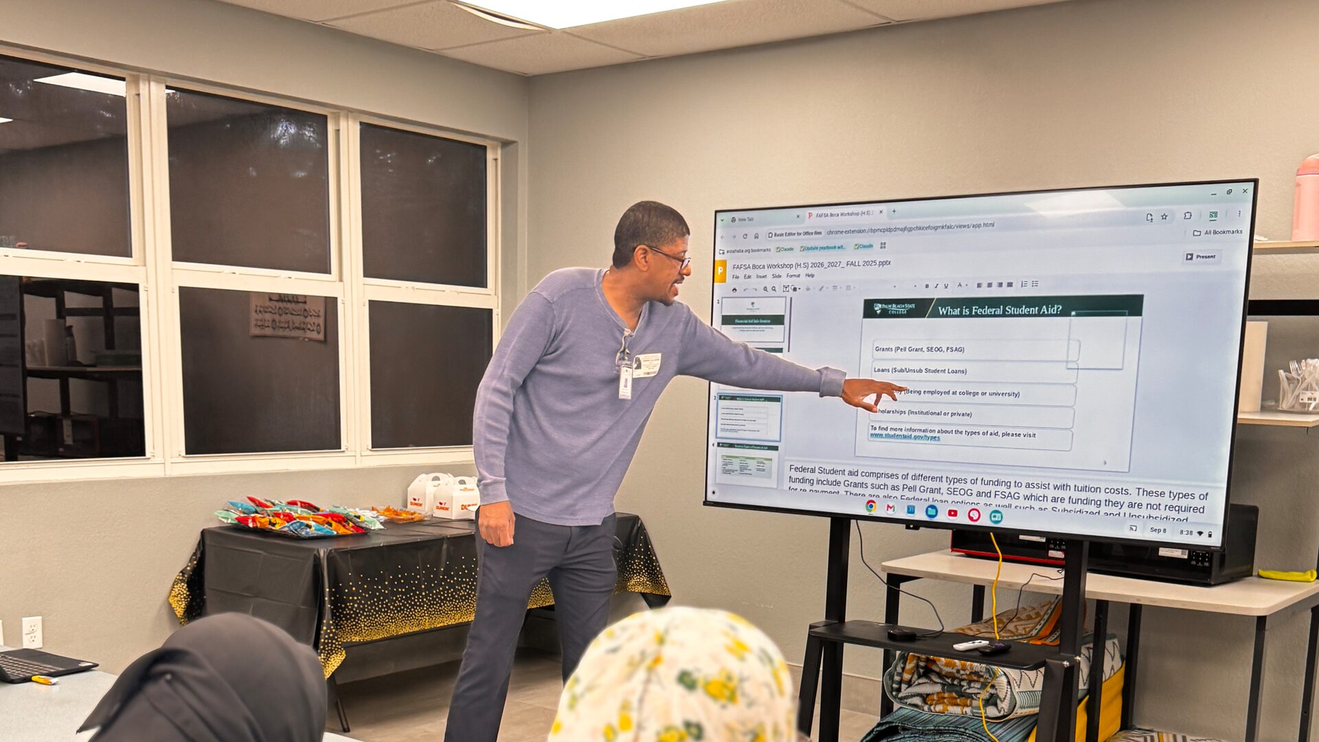 A presenter points to a slide titled “What is Federal Student Aid?” on a large screen while two parents watch from a table.