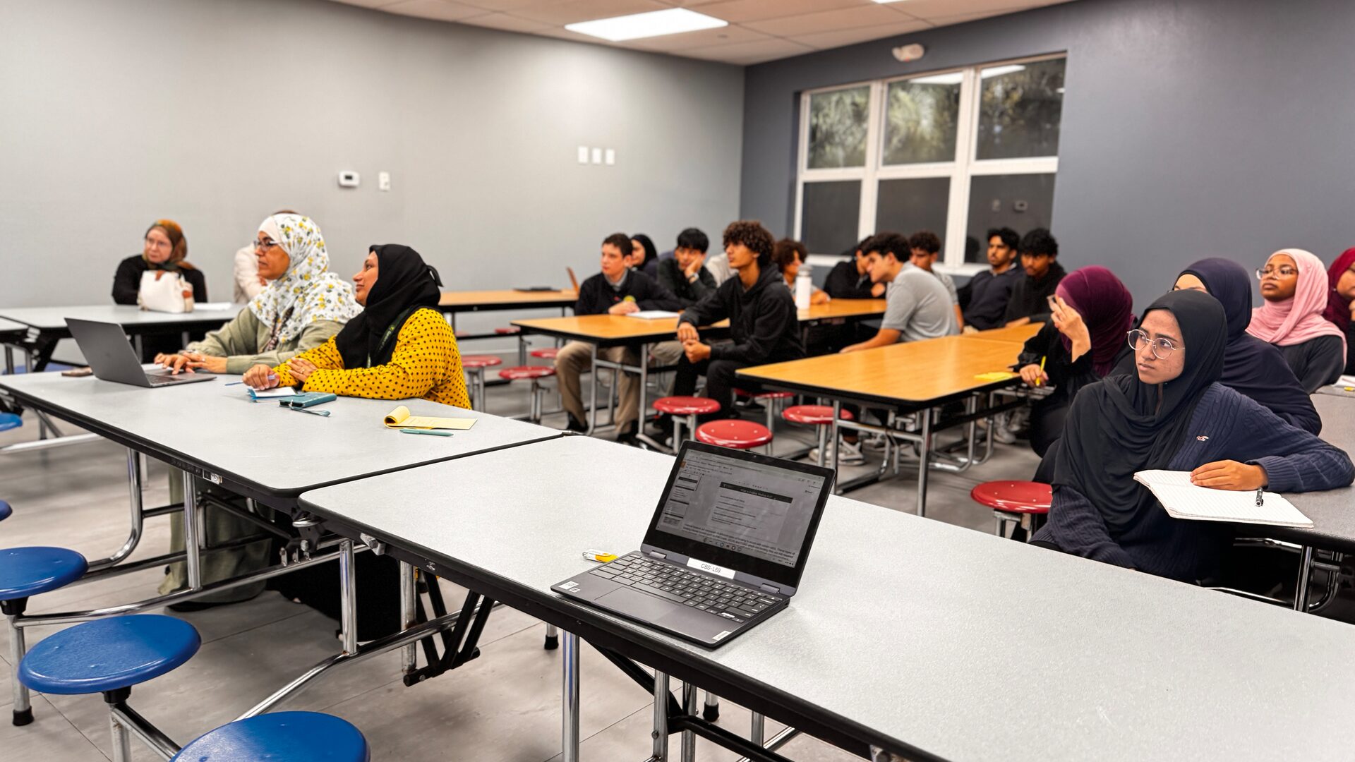 Students and parents sit at cafeteria tables, listening to the presentation.