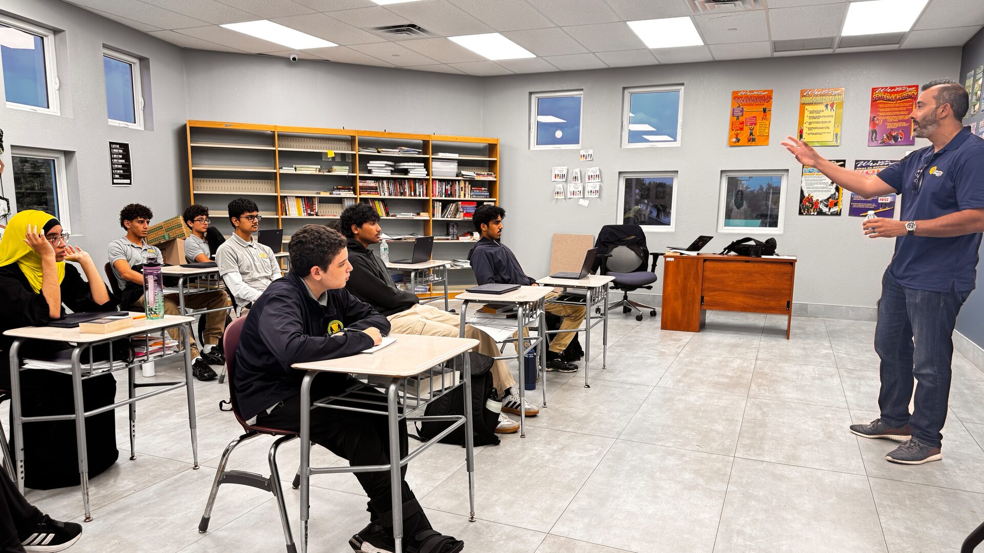 A guest speaker gestures as he talks to students seated at classroom desks.