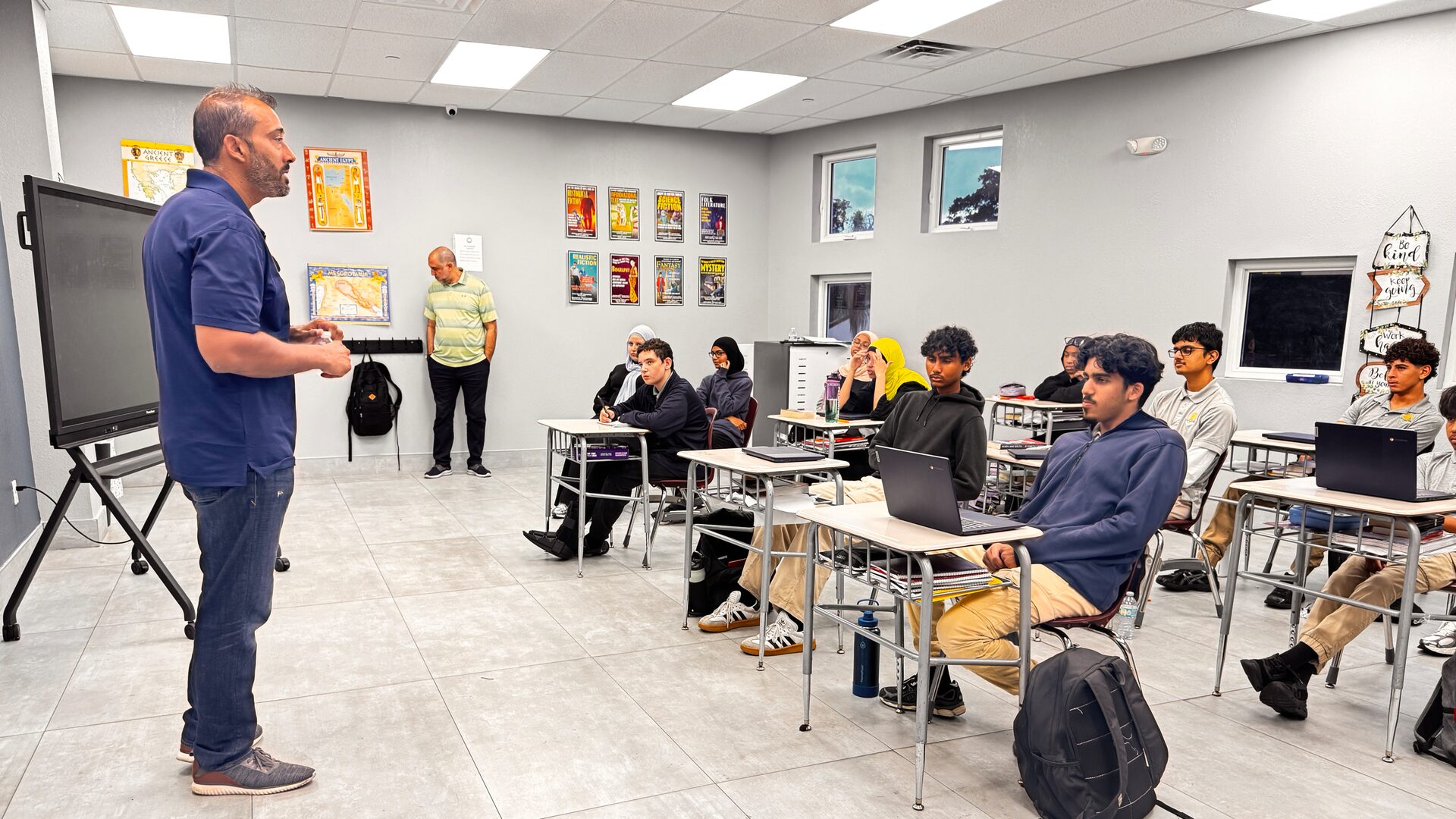 A guest speaker stands at the front of a classroom, speaking to students seated at desks with laptops.