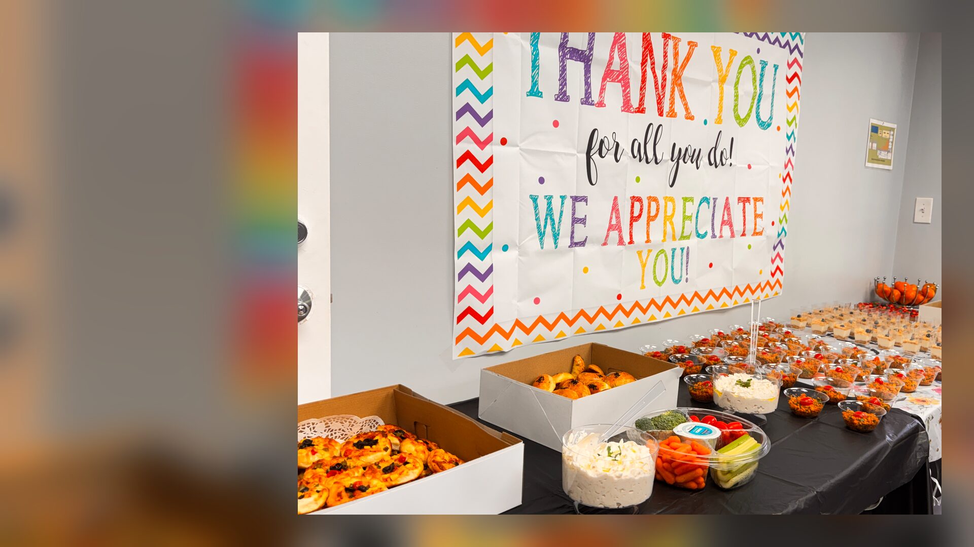 A banner reading “Thank you for all you do! We appreciate you!” hangs above a table of breakfast food.