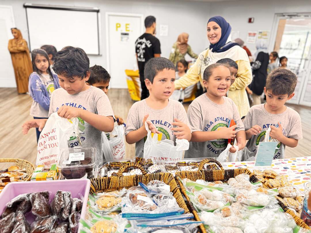 Young students fill their bags at a table of cookies and brownies as a volunteer smiles nearby