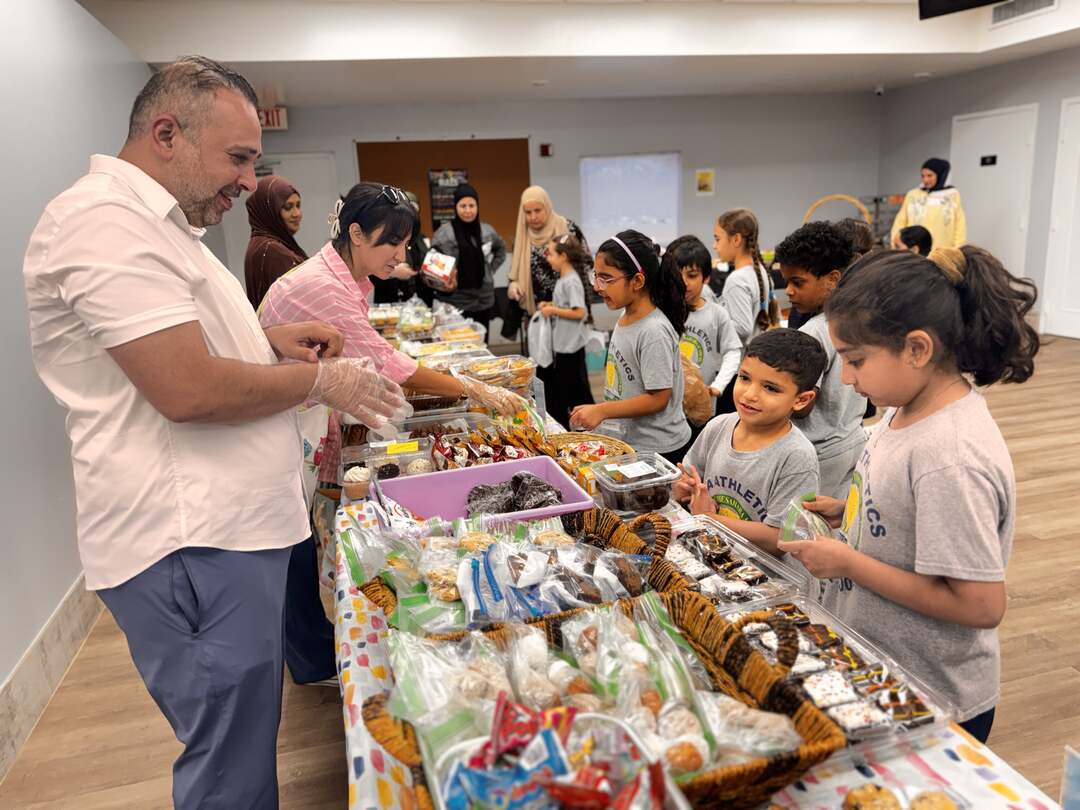 A gloved volunteer helps young students at a long table of sweets
