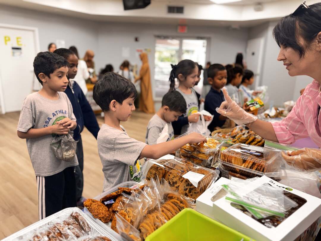 A smiling volunteer in gloves helps students choose cookies and pastries