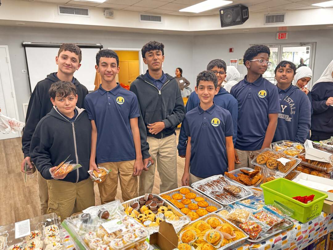 A group of older students pauses for a photo in front of trays of muffins and cookies, treats in hand