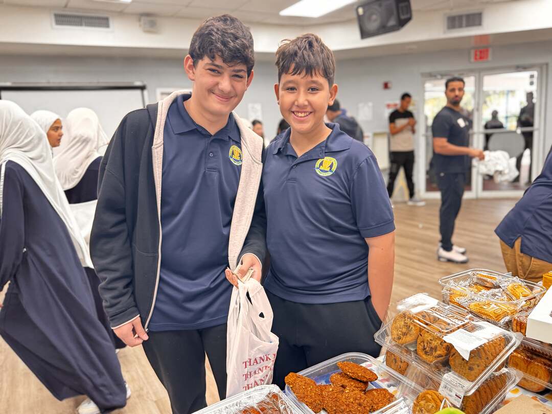 Two smiling students stand by a table of cookies with their bake sale bag