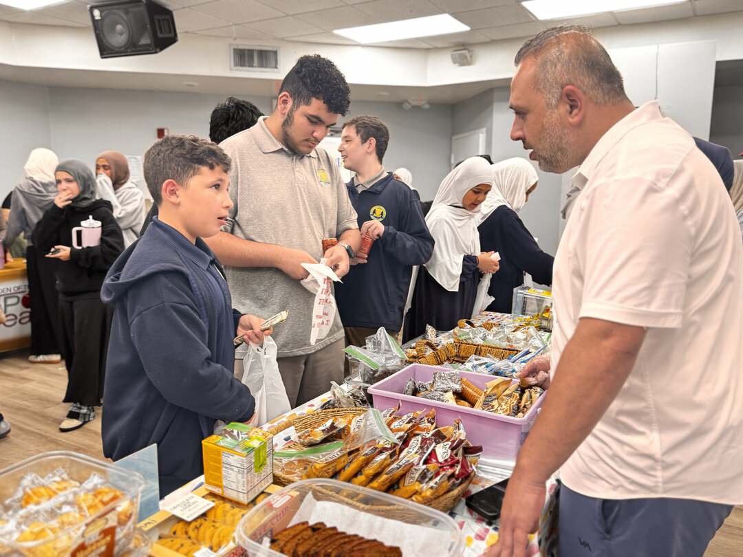 A volunteer helps an older student at a table of snacks and baked goods
