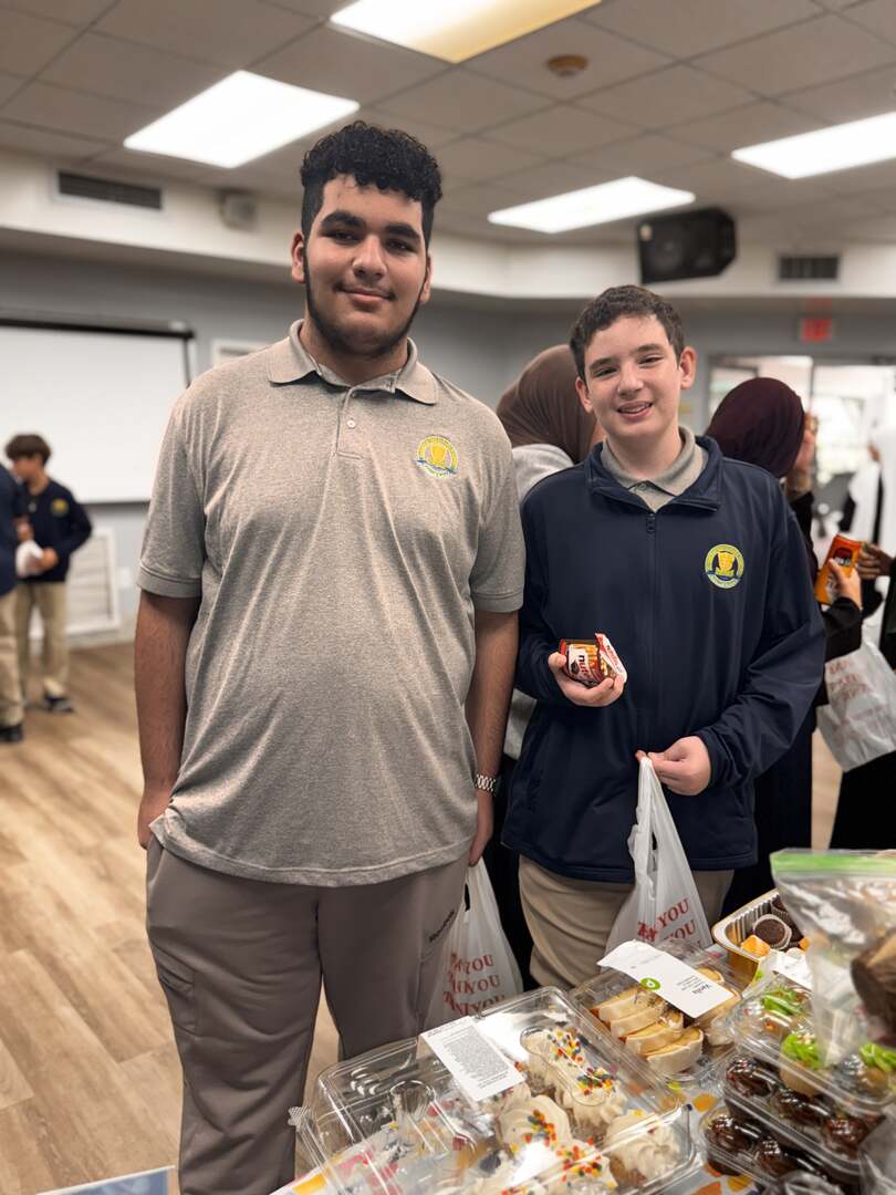 Two older students smile beside a table of frosted cupcakes, one holding a treat