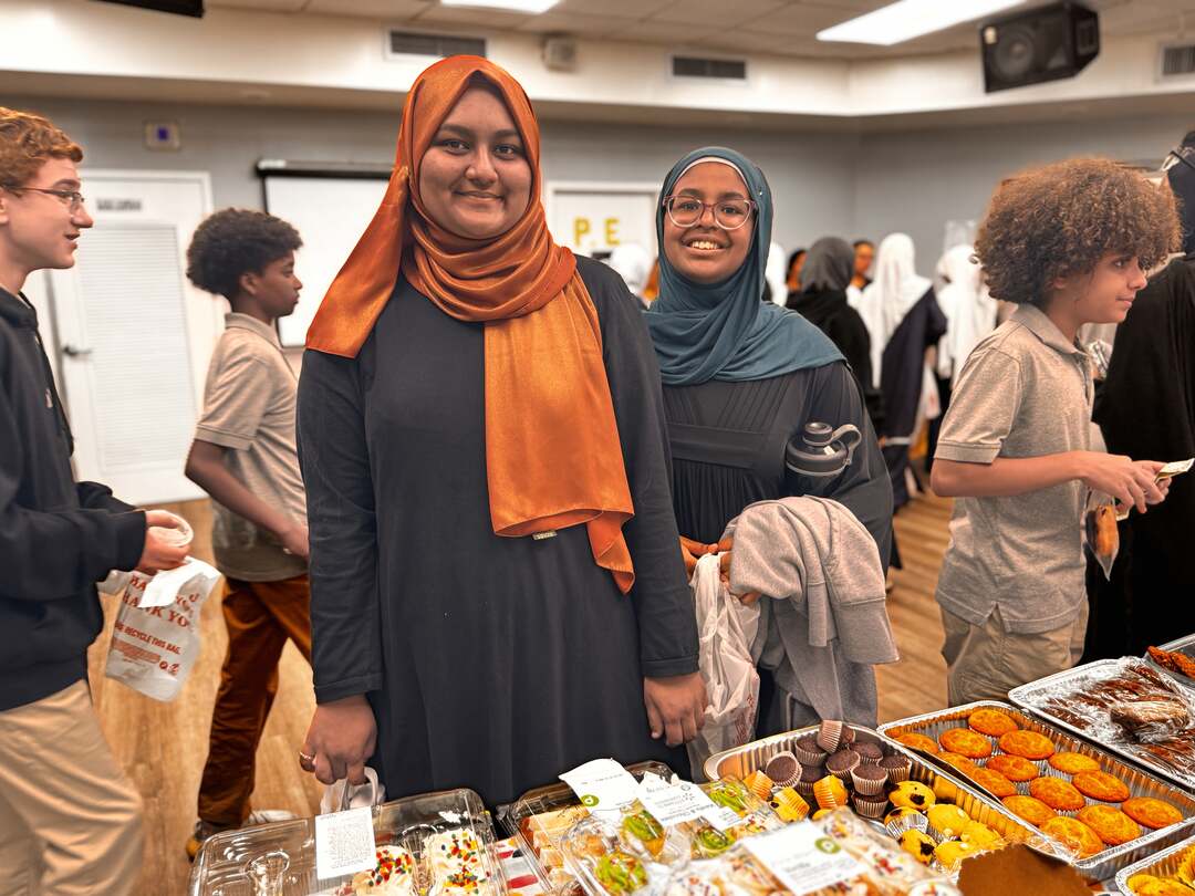 Two smiling shoppers pause behind trays of muffins and cupcakes