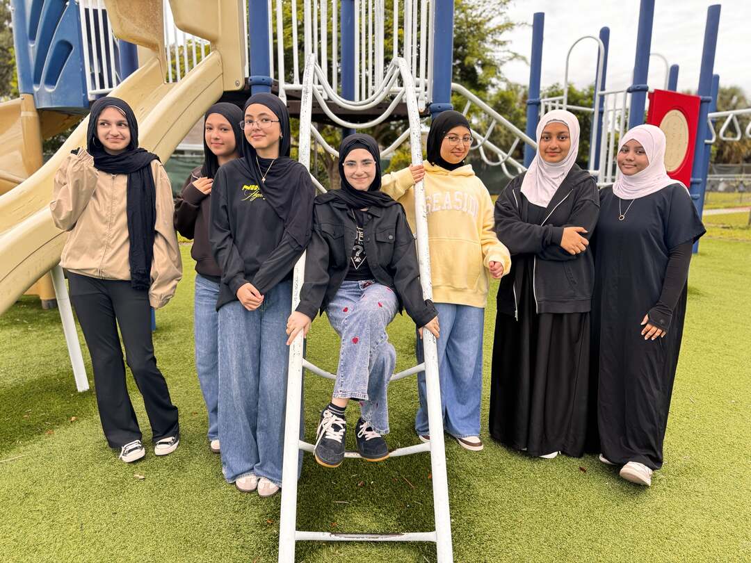 Friends in cozy hoodies and long dark dresses line up by the playground ladder on Dress Down Day