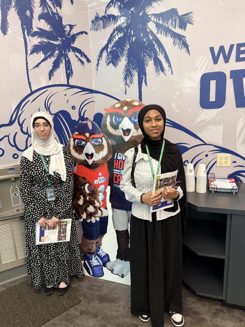 Two visitors with FAU brochures beside a cut-out of the owl mascot